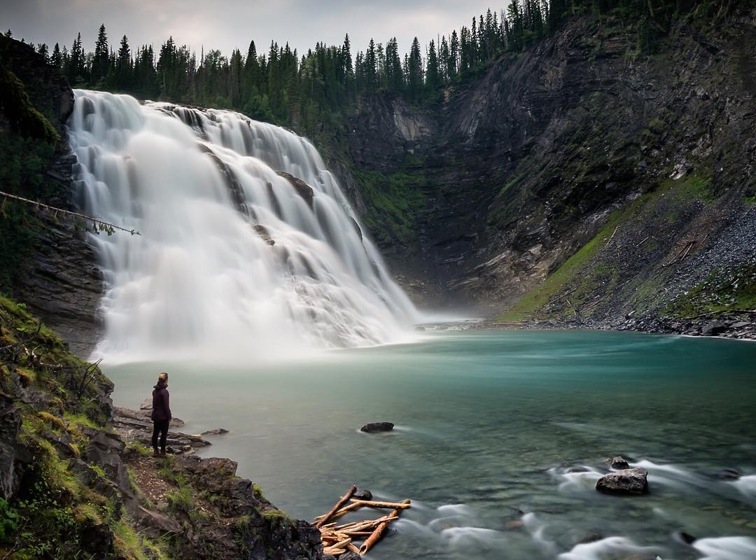 A wide and powerful waterfall tumbles down toward a clear blue pool, surrounded by forest.