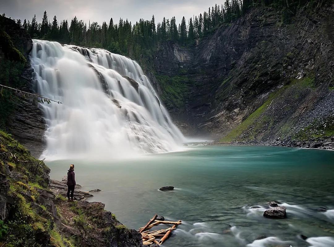 A wide and powerful waterfall tumbles down toward a clear blue pool, surrounded by forest.