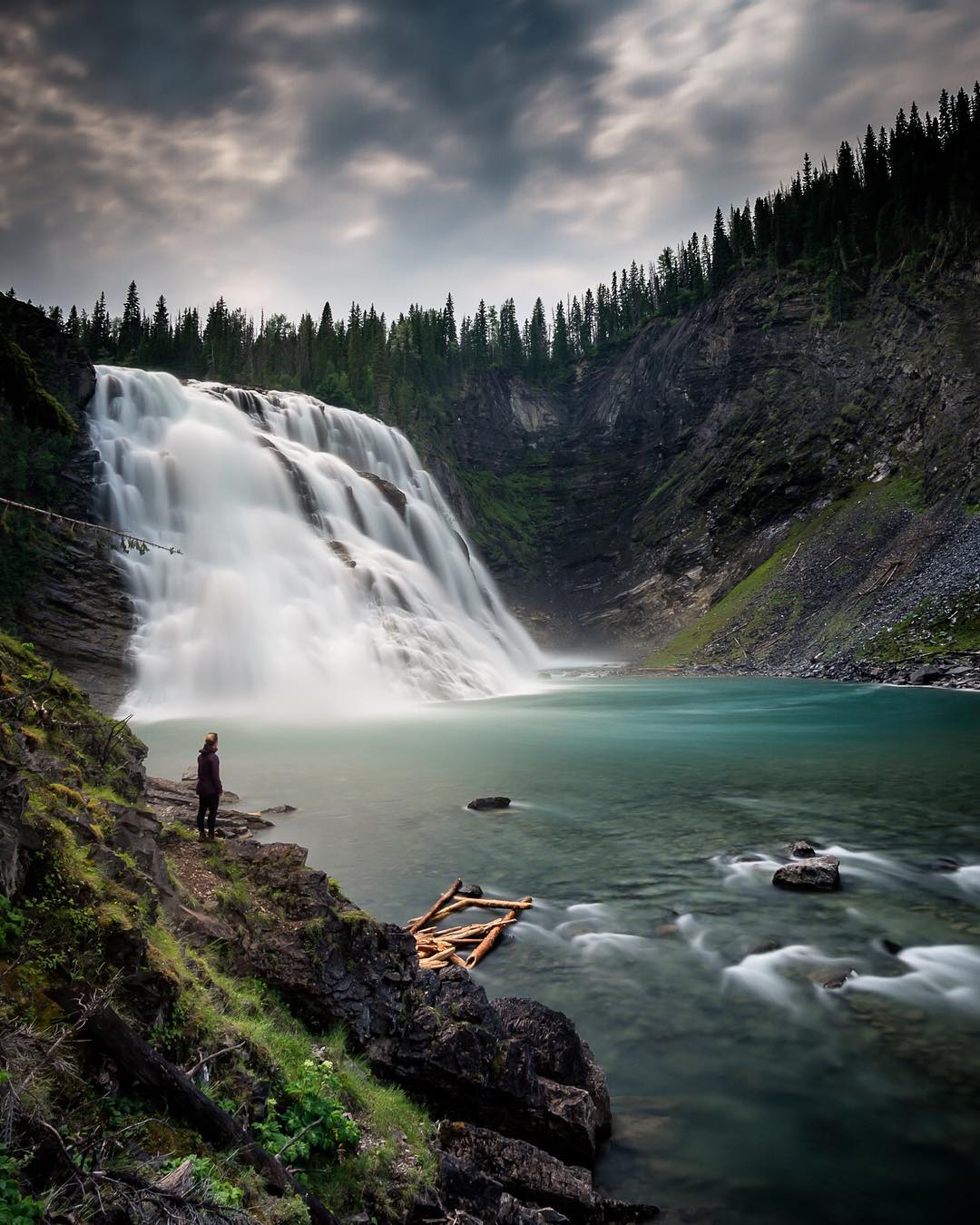 A wide and powerful waterfall tumbles down toward a clear blue pool, surrounded by forest.