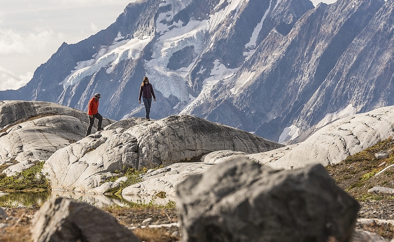 Two hikers walk across giant boulders with a mountain in the background