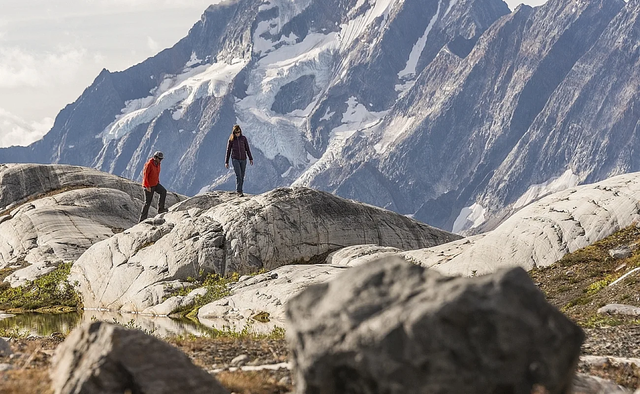 Two hikers walk across giant boulders with a mountain in the background