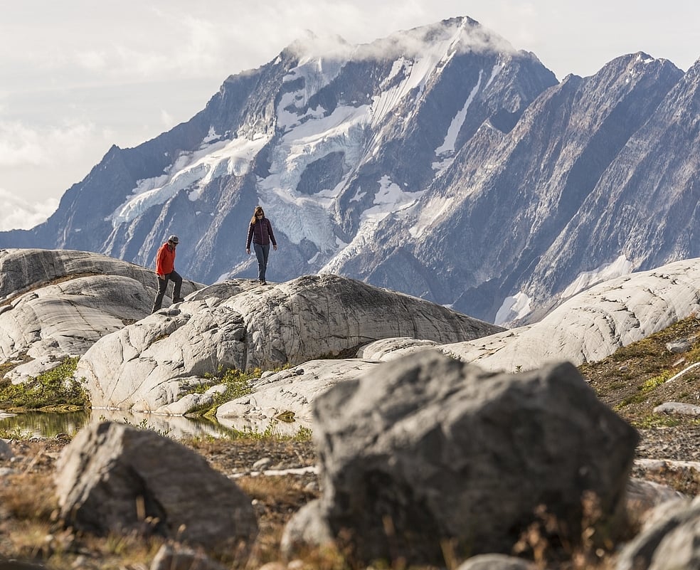 Two hikers walk across giant boulders with a mountain in the background