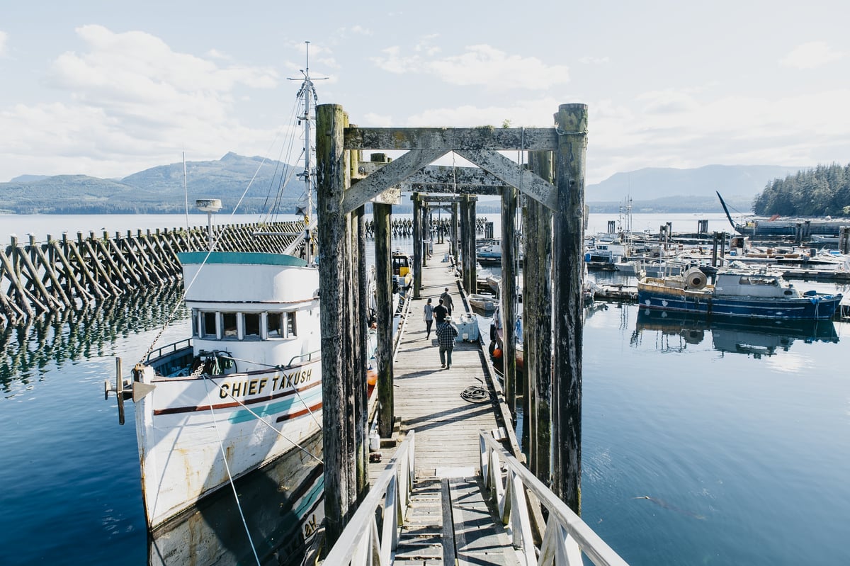 A wooden dock with one boat moored in the foreground and several others farther out on the right. In the distance is the forested mountains of Vancouver Island.