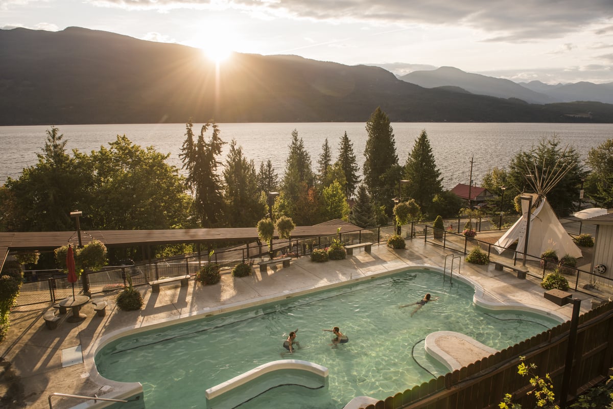 Bathers in an outdoor pool at Ainsworth Hot Springs during the daytime. The sun is starting to set behind the mountain and a lake is behind the pool.