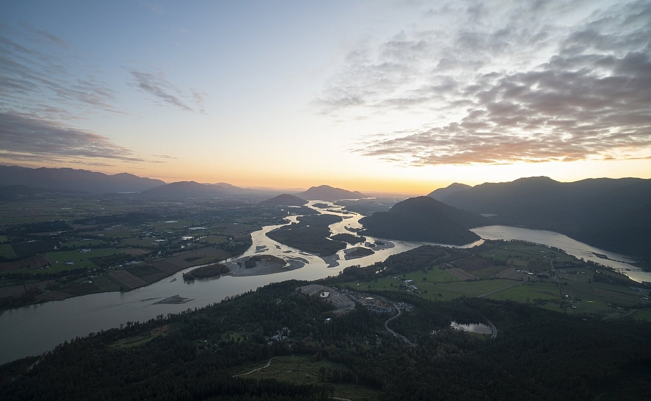 A sweeping view of the Fraser River on a clear day. There are silhouettes of mountains in the backdrop crowned by the setting sun.