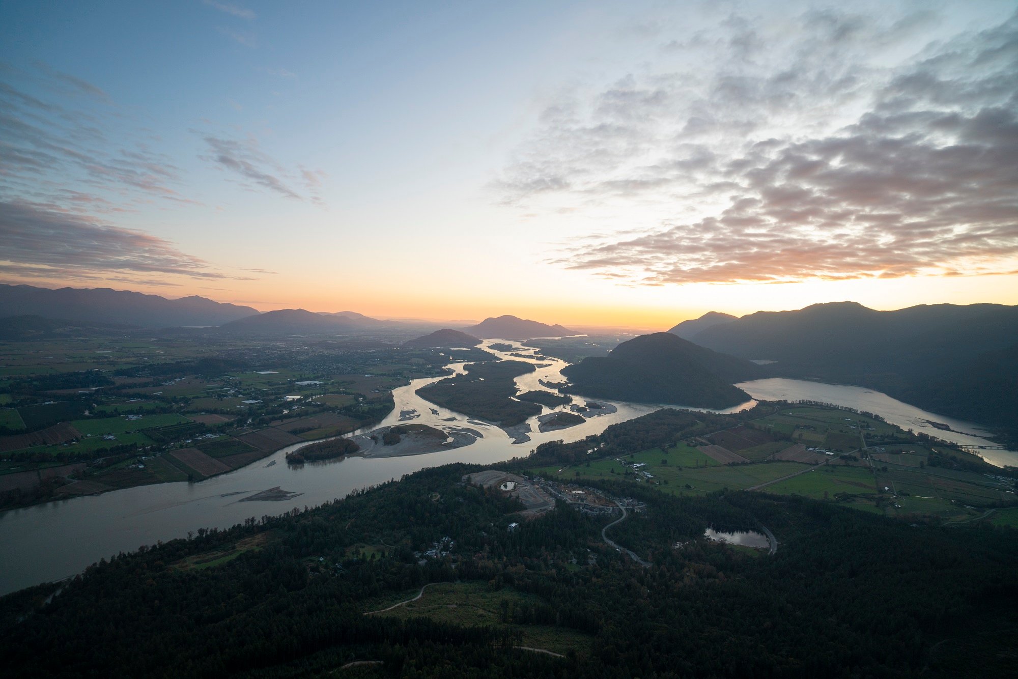 A sweeping view of the Fraser River on a clear day. There are silhouettes of mountains in the backdrop crowned by the setting sun.