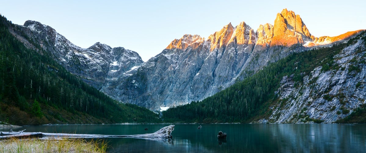 Rugged snow capped mountains frame a lake in Strathcona Provincial Park. The sun hits the top of the farthest mountain and the lake below is in shadows.