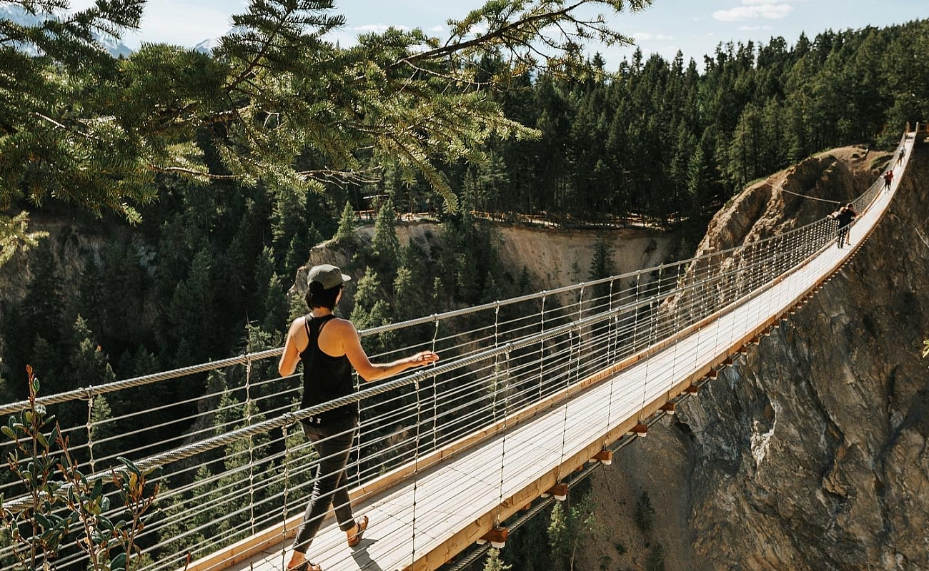 A woman wearing a black tank top, black pants and a hat walks across a suspension bridge on a summer day.