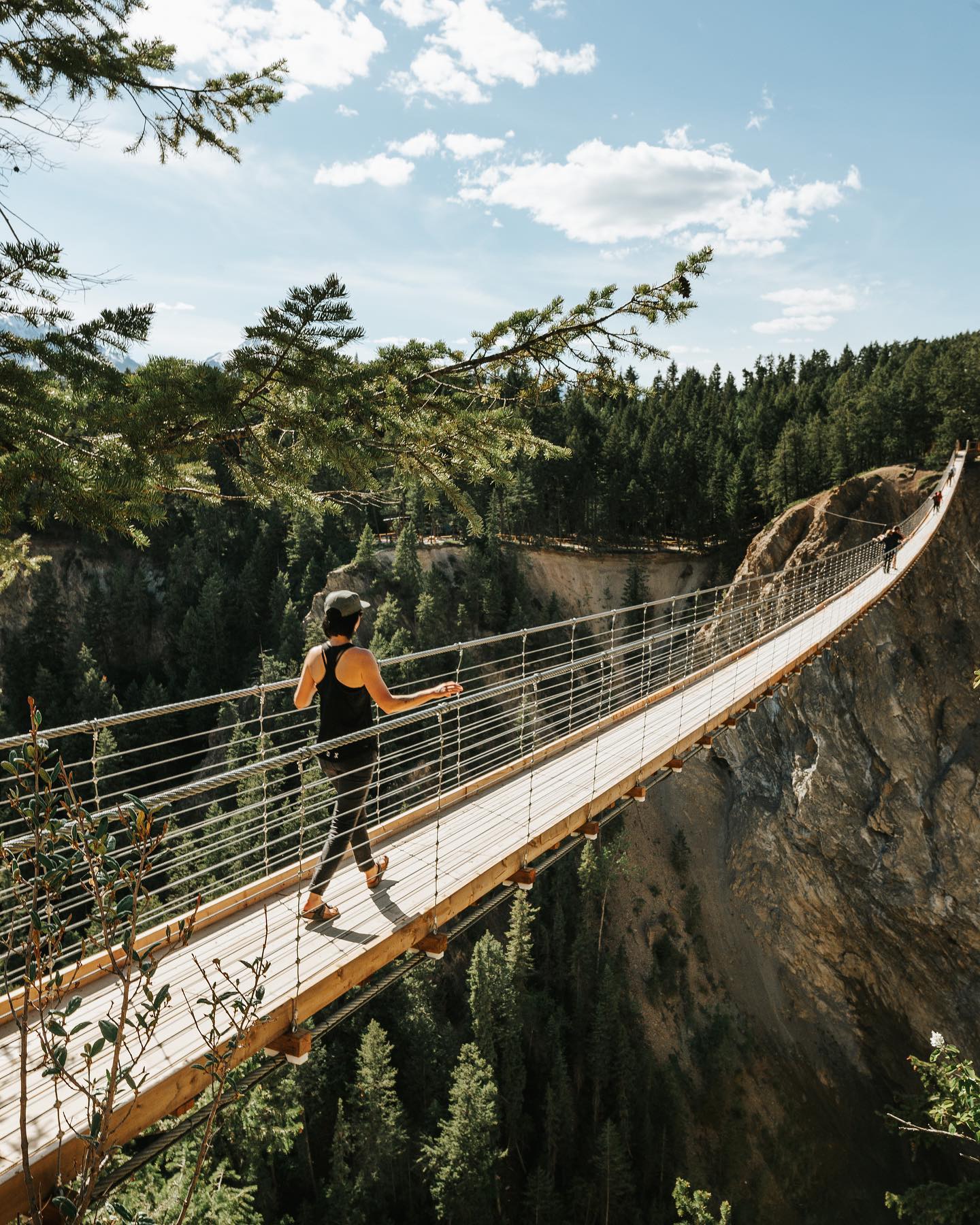 A woman wearing a black tank top, black pants and a hat walks across a suspension bridge on a summer day.