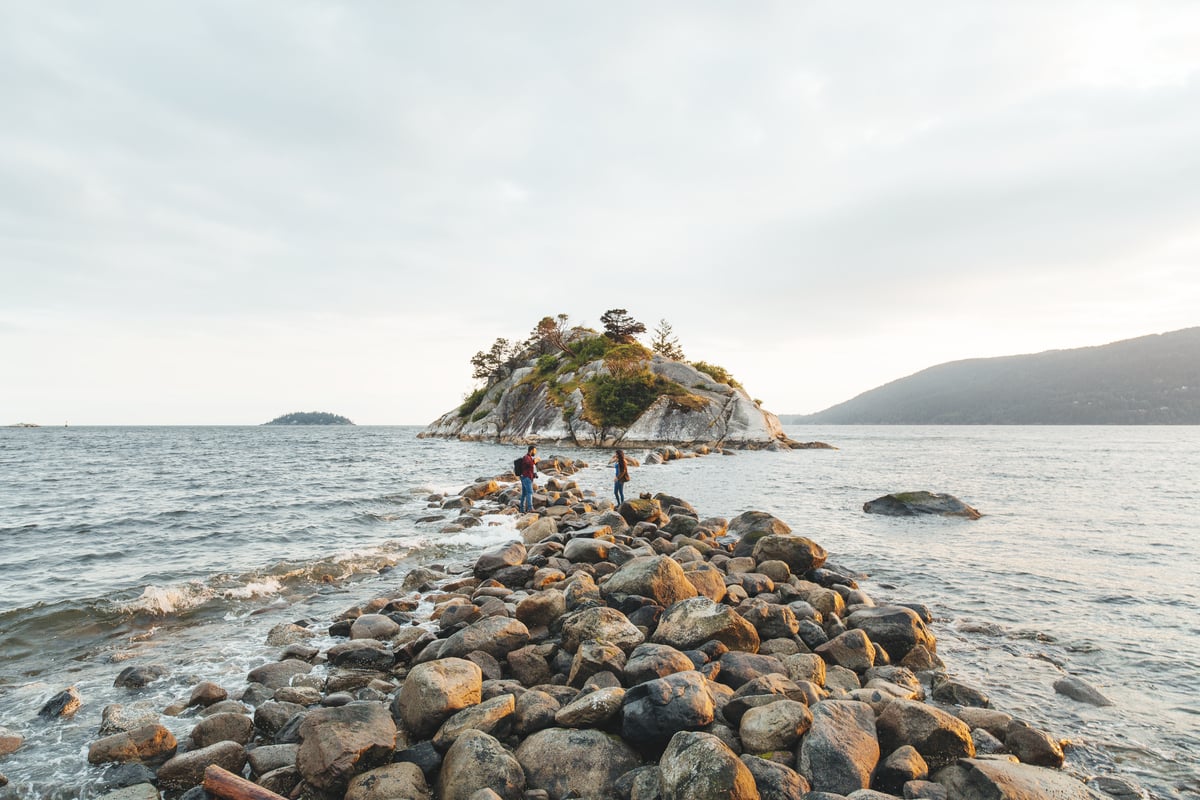People walking on rocks by the ocean in Whytecliff Park, West Vancouver. There is a small rocky island in front and clouds on an overcast day.