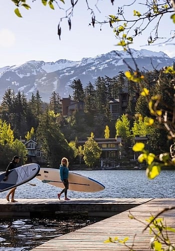 Zwei Personen tragen Paddleboards auf dem Steg eines Sees mit Häusern am Ufer dahinter.