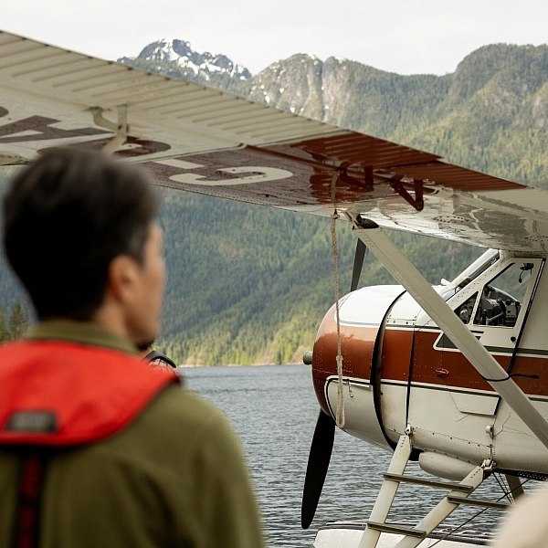 Zwei Menschen blicken auf ein Wasserflugzeug auf einem von Bergen umgebenen Gewässer.
