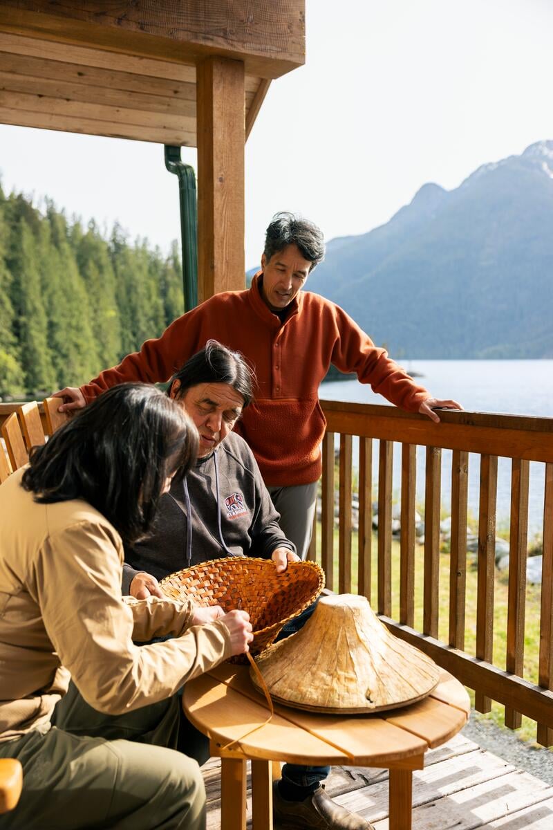 Three people are on a deck overlooking an inlet. One is an Indigenous guide showing the other two how to weave a cedar hat.