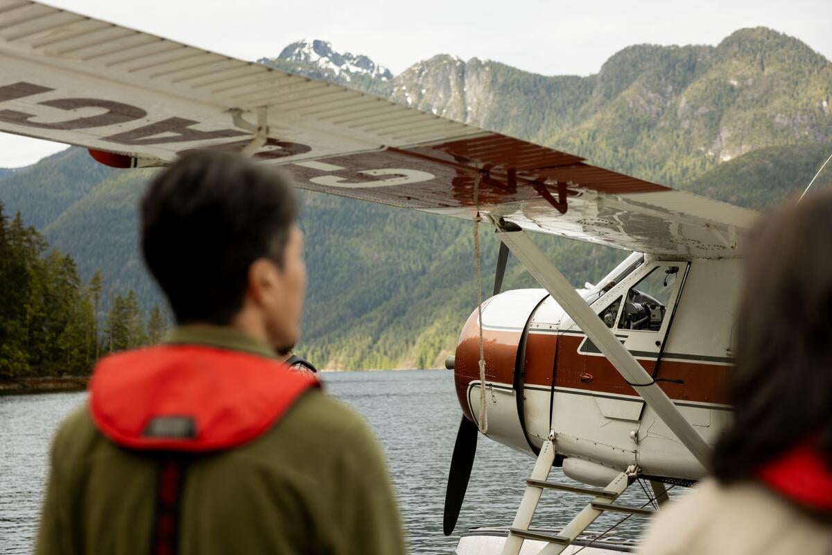 Two people look toward a seaplane on a waterway backed by mountains.
