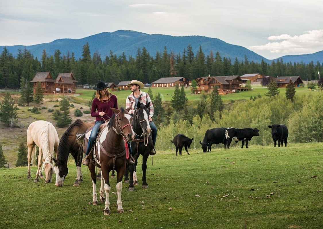 Zwei Personen zu Pferd auf einem Feld mit anderen Pferden und Kühen, im Hintergrund eine weitläufige Ranch.