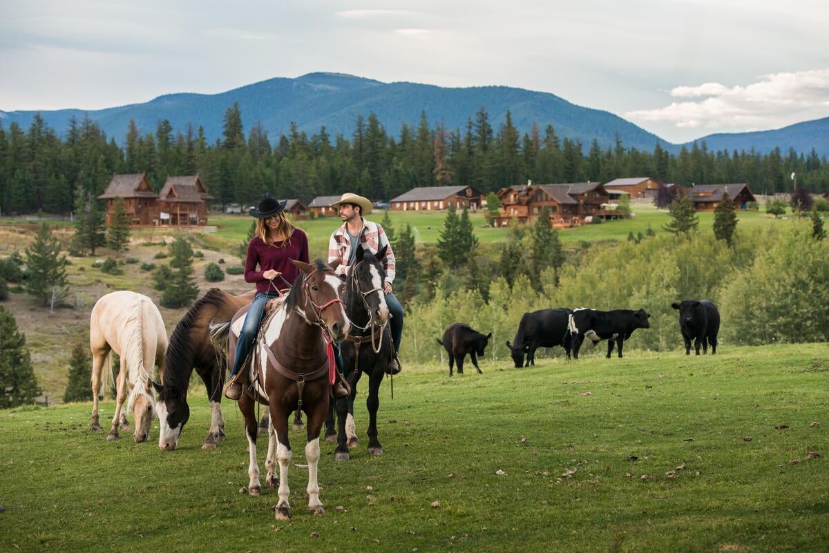 Two people on horseback in a field with other horses and cows with a sprawling ranch in the background.