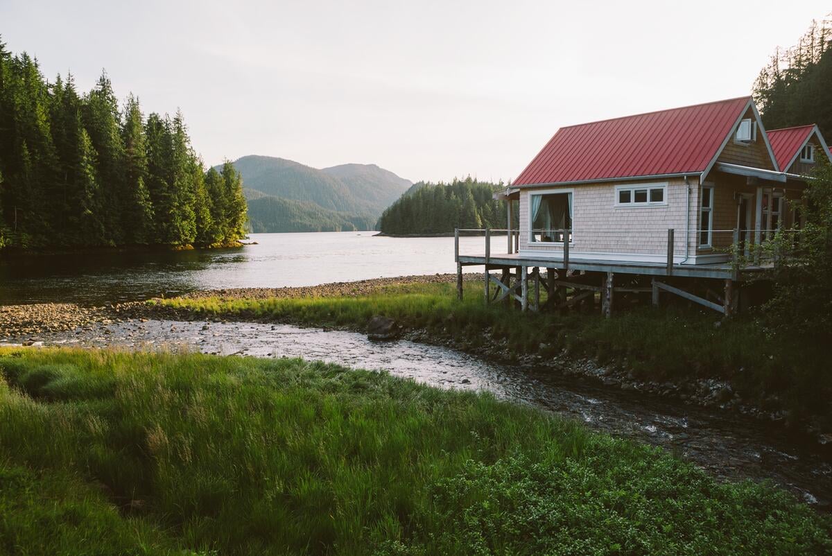 Two lodge building sit on a raised platform on the banks of an inlet.