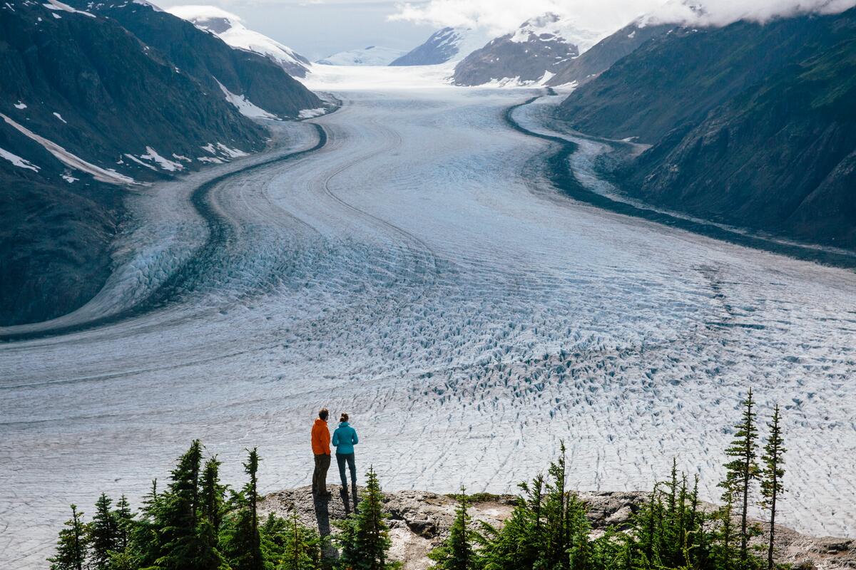 Two people stand on a bluff overlooking a massive glacier.