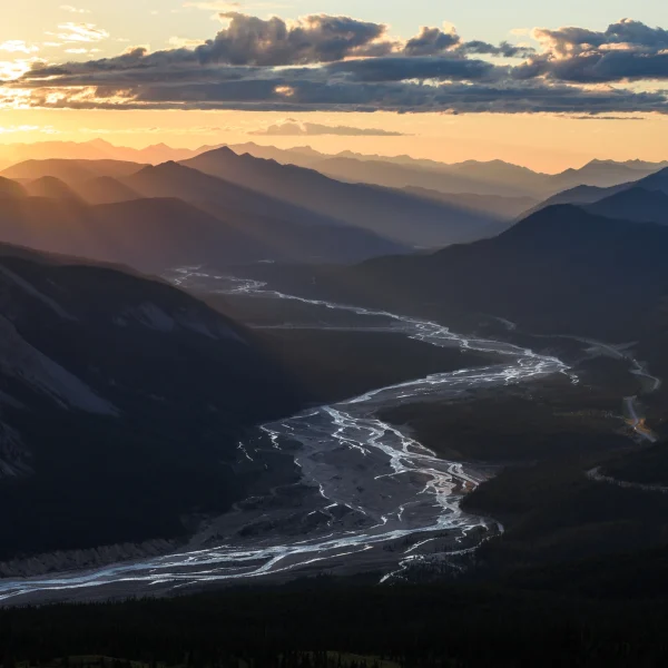 Sonnenuntergang über einem Bergtal mit einem Fluss, der sich durch die Mitte schlängelt, während Lichtstrahlen über die Gipfel tanzen.
