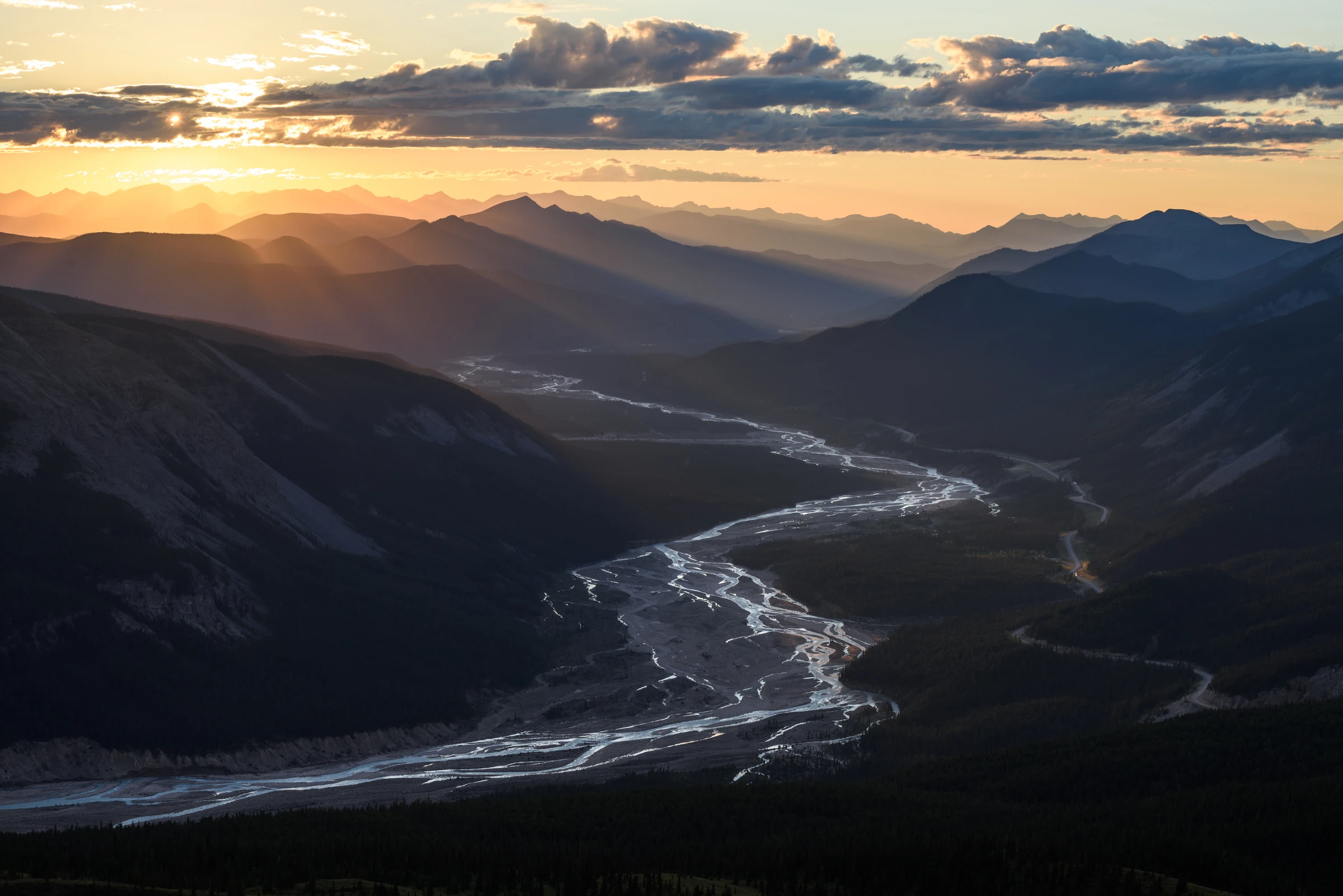 Sunset over a mountain valley with a river slicing through the centre as rays of light dance across the peaks.