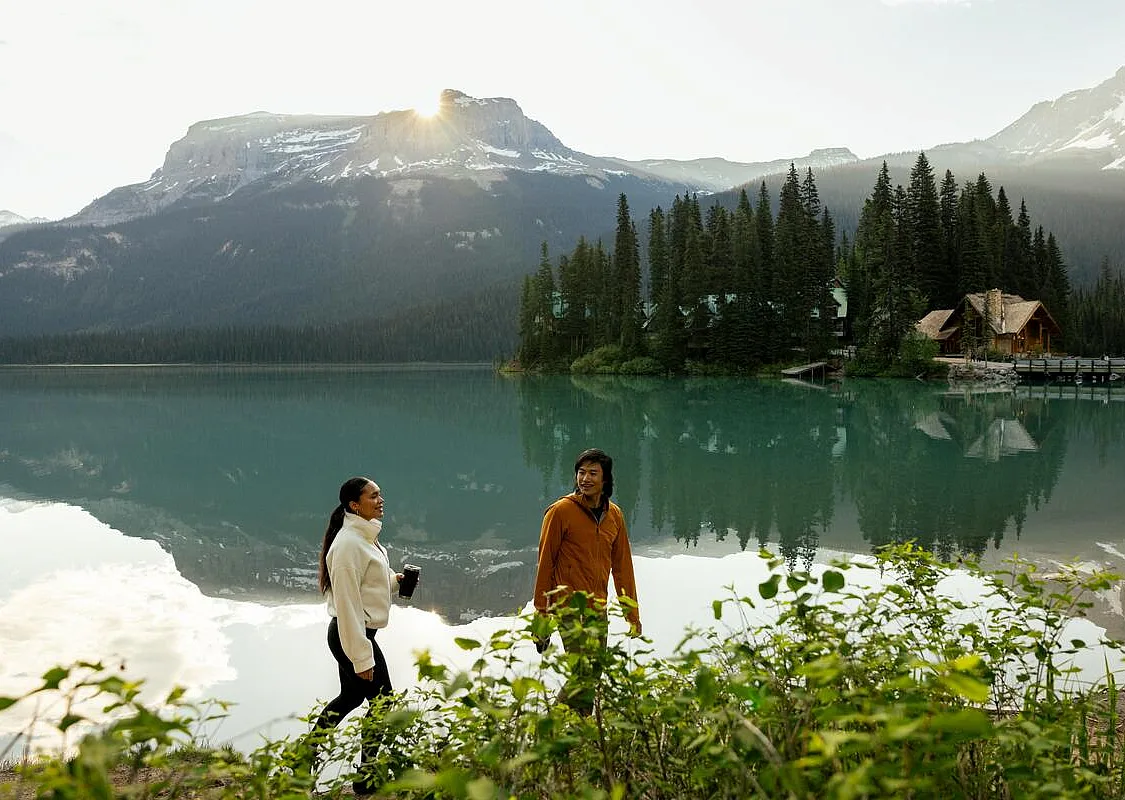 Dos amigos paseando por la orilla del lago Emerald, en el Parque Nacional Yoho, en las Montañas Rocosas canadienses, con el Emerald Lake Lodge al fondo.