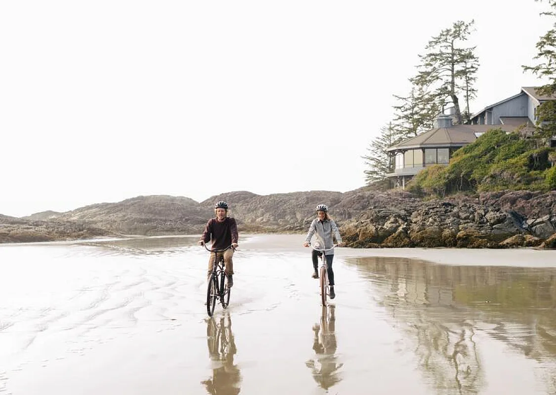 Dos personas montando en bicicleta por North Chesterman Beach, en Tofino, con el Wickaninnish Inn al fondo.