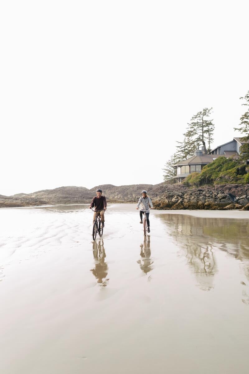 Two people riding bikes along North Chesterman Beach in Tofino, with the Wickaninnish Inn in the background..