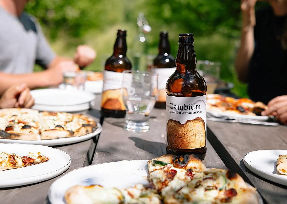 Primer plano de una botella de sidra de Cambium Cider Co. en una mesa de pícnic con pizza, gente y más sidra de fondo.